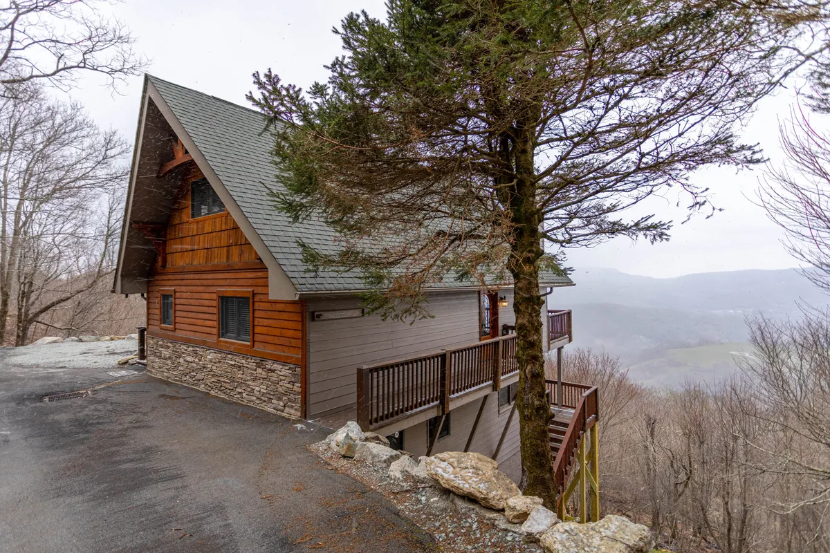 A wide view of the Grand Panorama cabin nestled among tall trees, with the cabin's rustic structure set against a backdrop of distant mountain peaks. The porch invites relaxation while the surrounding forest hints at privacy and seclusion.