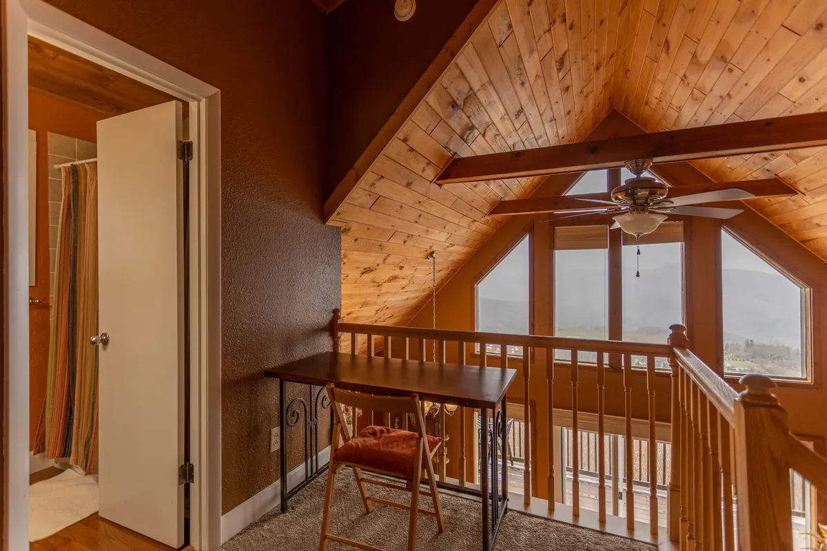 A serene interior space with a wooden ceiling, large windows overlooking the landscape, and a small table with a chair. The room is bathed in tranquil light, offering views outward and a peaceful corner for reading or quiet reflection.