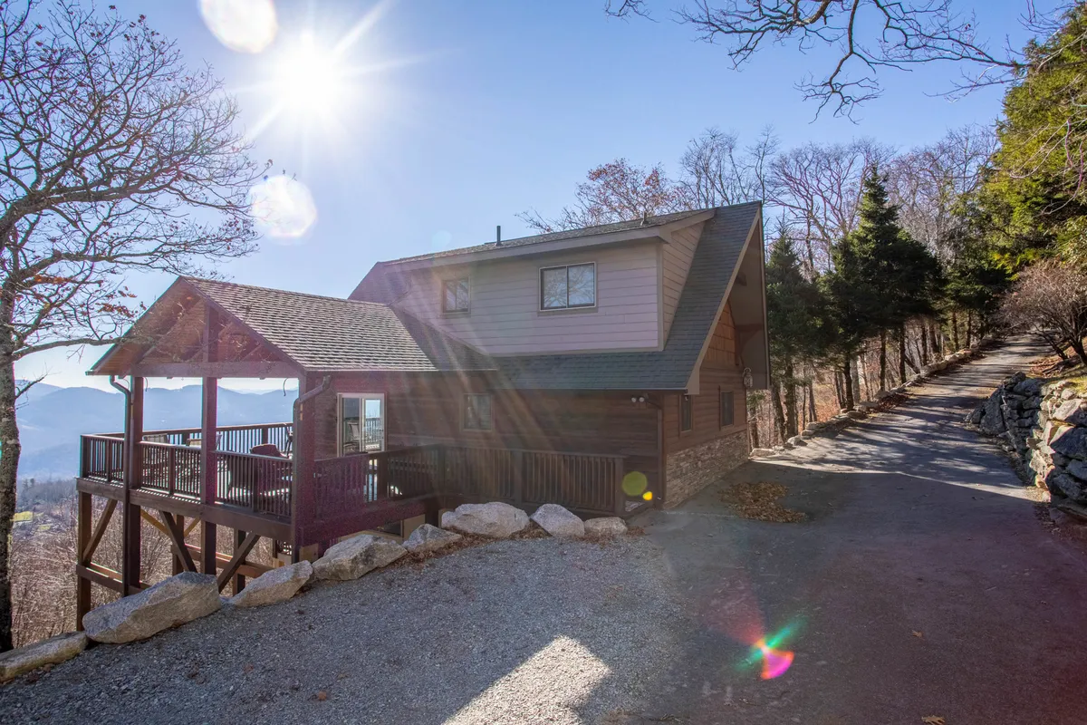 This image presents the house exterior and spacious deck surrounded by lush trees, with panoramic mountain views stretching in the background. The deck is arranged for outdoor relaxation and socializing, evoking a sense of peaceful retreat in nature.