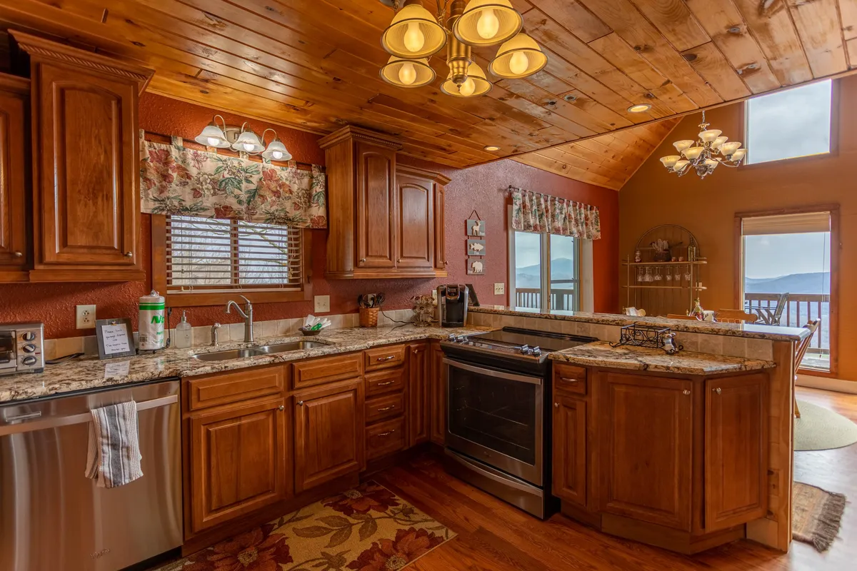 A bright and welcoming kitchen space featuring granite countertops, wood cabinetry, a spacious island, and large windows that let in plenty of natural light. The scene invites cooking, socializing, and enjoying coffee with a mountain sunrise view.